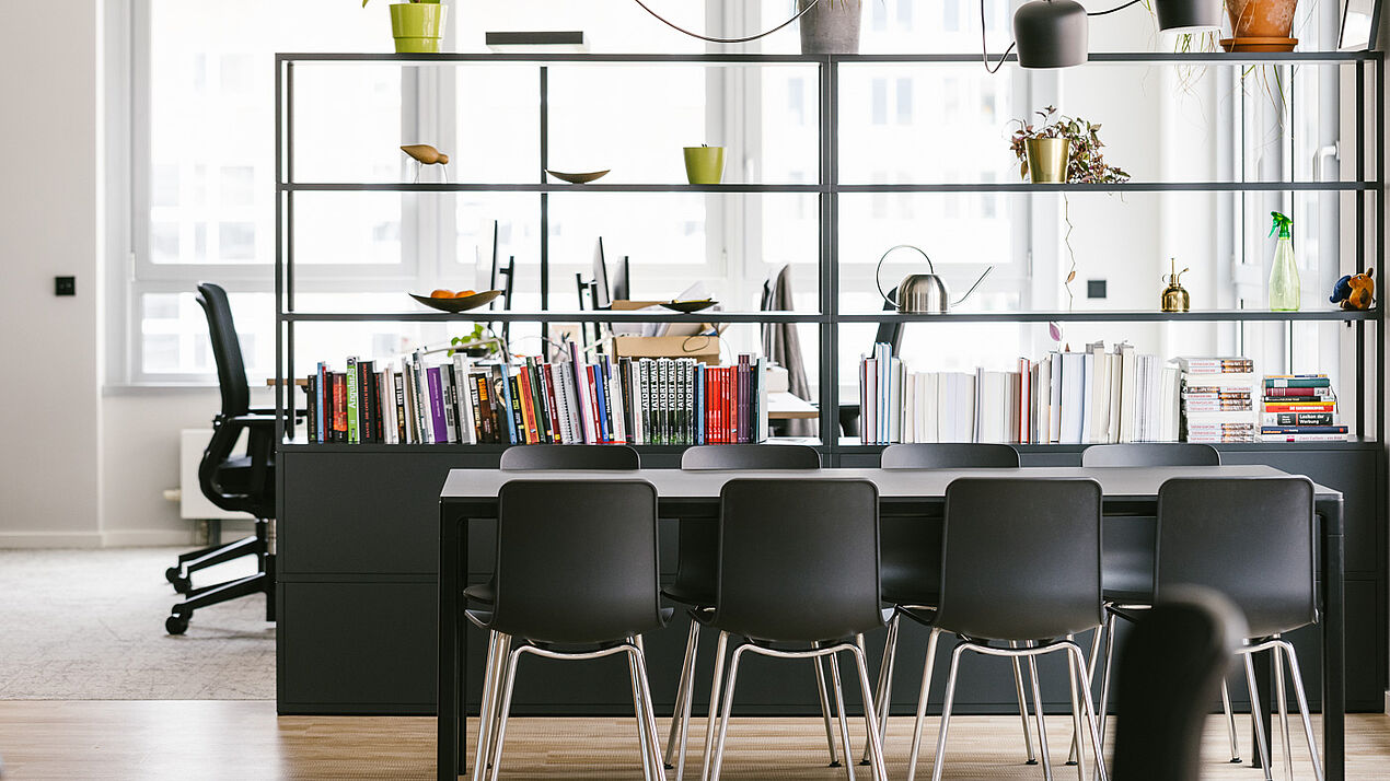 Offener Arbeits- und Besprechungsbereich mit langem Tisch, mehreren Stühlen, offenem Regal mit Büchern und Pflanzen sowie großen Fenstern in modernem Bürodesign.