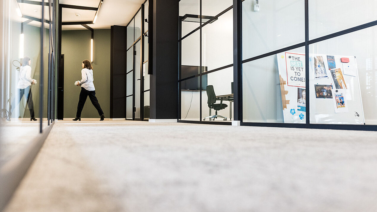 Moderne Büroflure mit Glaswänden und Besprechungsräumen, eine Person geht durch den Gang, im Hintergrund sind Arbeitsplätze und eine Pinnwand sichtbar.