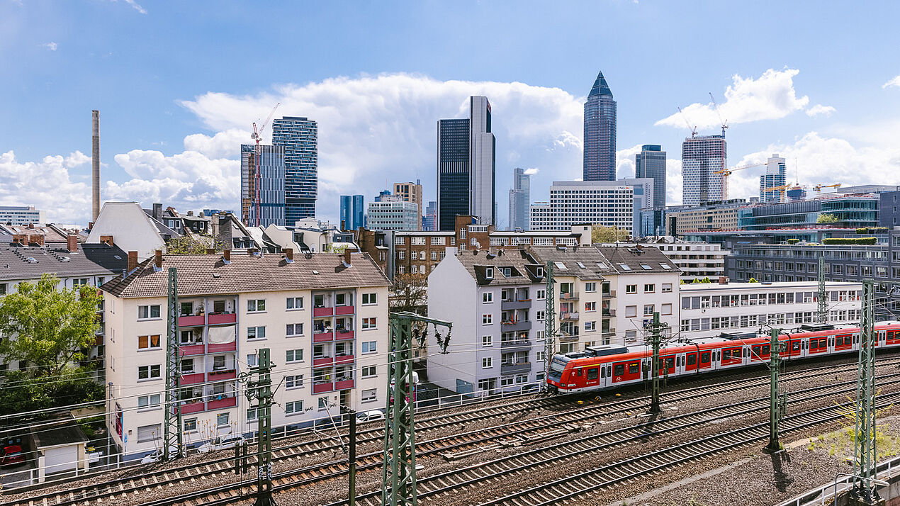 Ansicht von Frankfurt mit Wohnhäusern im Vordergrund, Bahntrassen mit roter S-Bahn und der Frankfurter Skyline mit Hochhäusern unter blauem Himmel.
