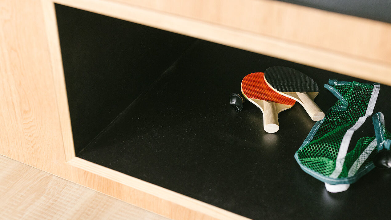 Offenes Regalfach mit Tischtennisschlägern, Ball und Netz, aufgeräumt in einem hellen Holzmöbel in einem Bürobereich..